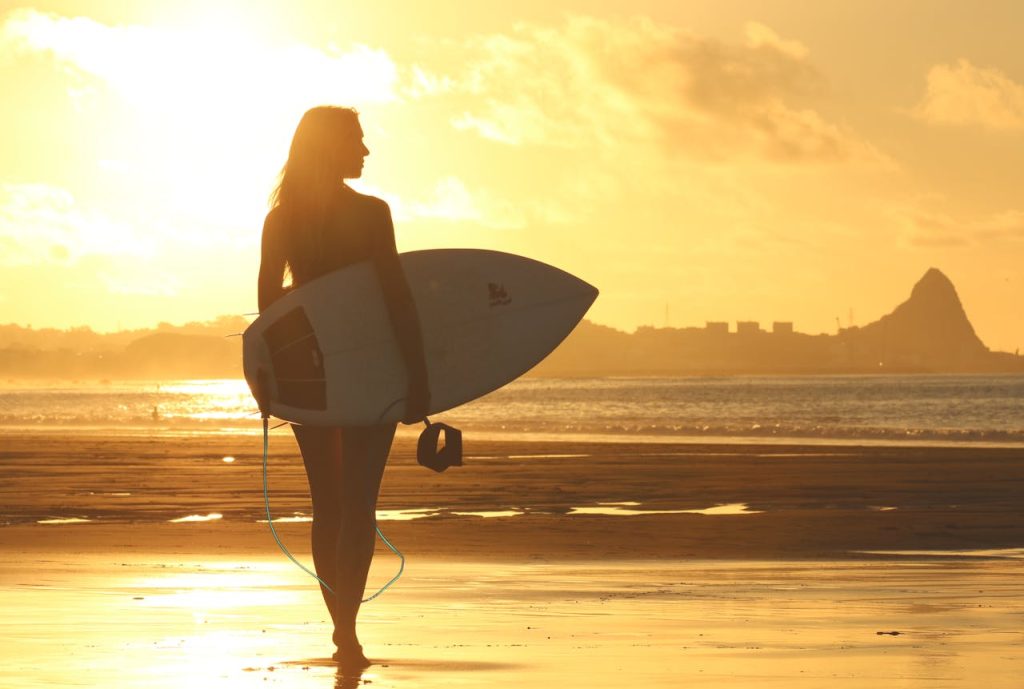A silhouette of a woman holding a surfboard, standing on a beach at sunset with a golden sky.