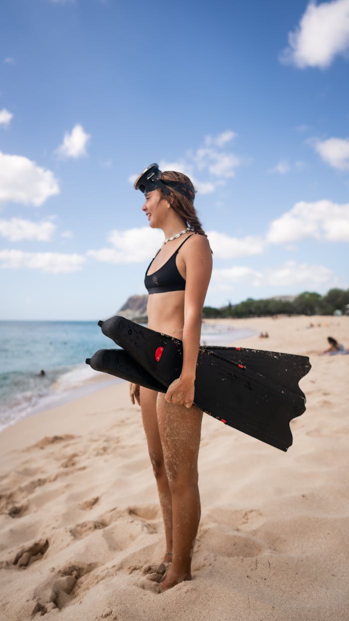 A young woman in swimwear holds snorkeling fins on a sunny tropical beach.